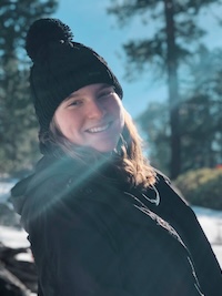 A headshot of Anna wearing a black knit beanie with one of those cute little poofs on it, in front of a snowy background.